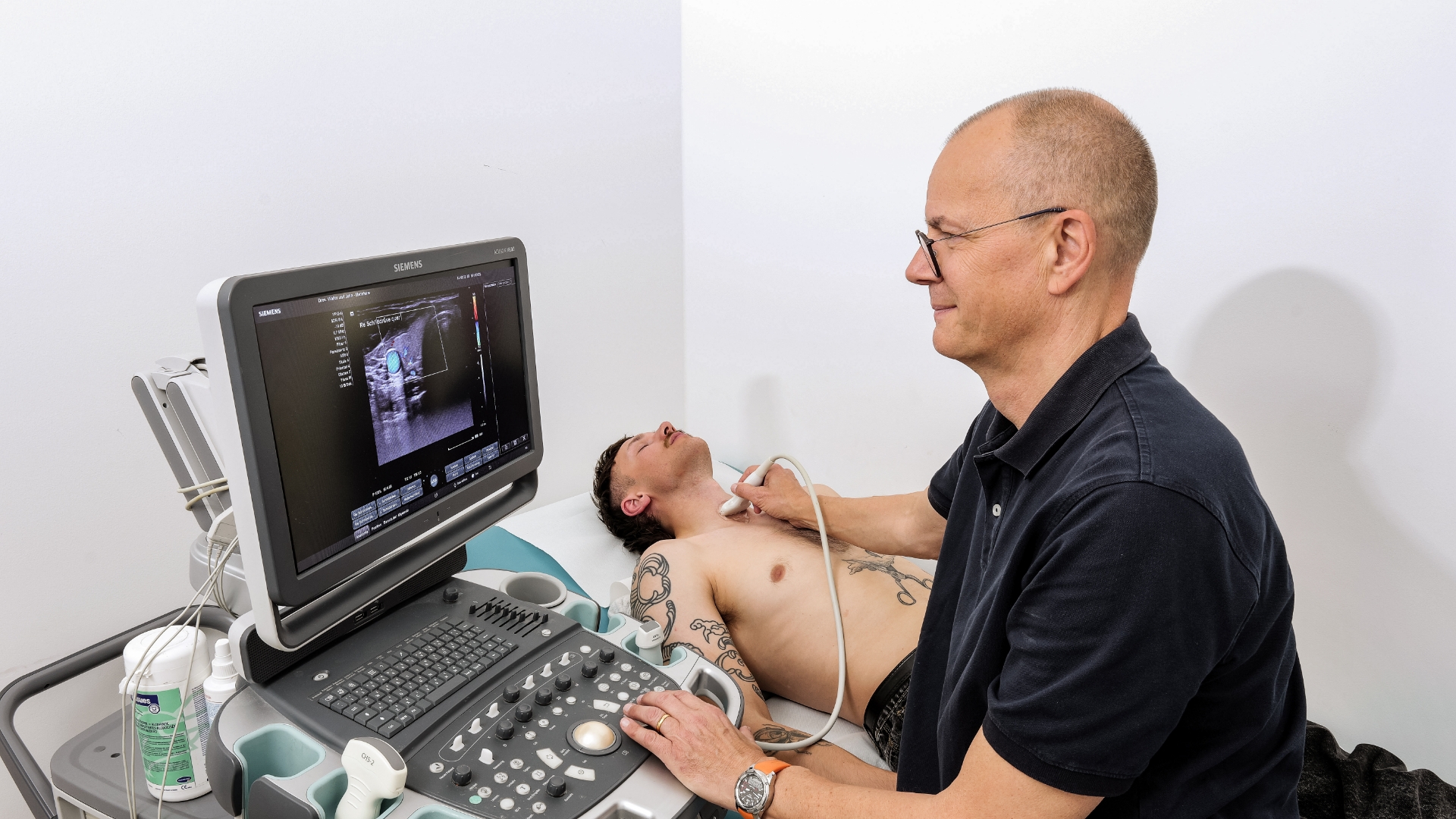Arzt nimmt Ultraschall der Halsschlagader eines jungen Mannes vor, der auf einer Pritsche liegt.